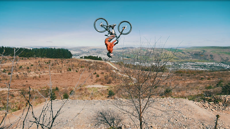 a bike rider does a backflip in the countryside. Hook and loop is used in his gear and apparel